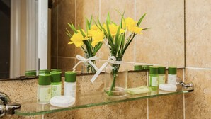 Casa de banho com prateleira de vidro com produtos de higiene e um vaso com flores amarelas.