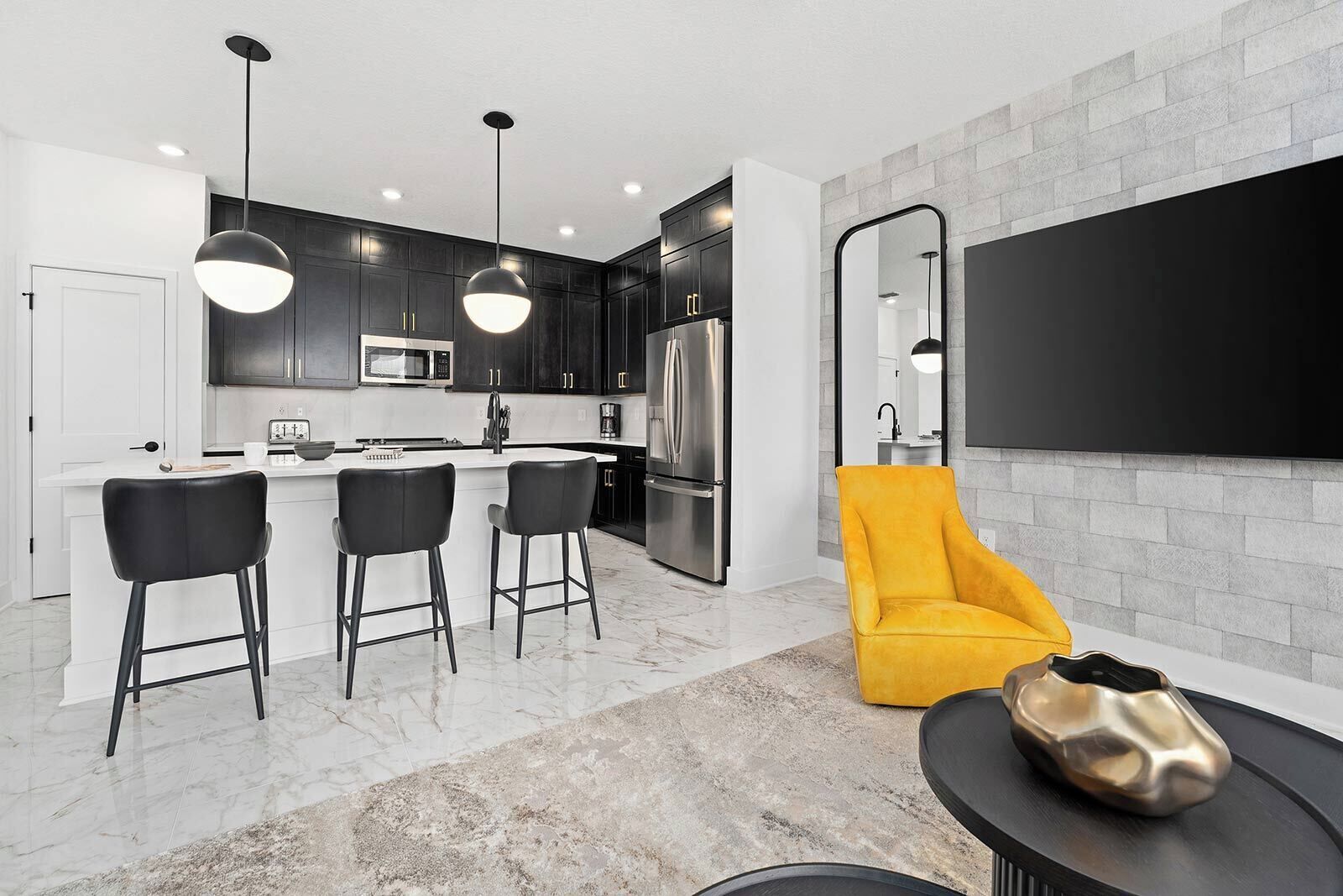 A modern kitchen with dark cabinets, a central island with bar stools, a large flat-screen TV mounted on the wall, and a yellow chair.
