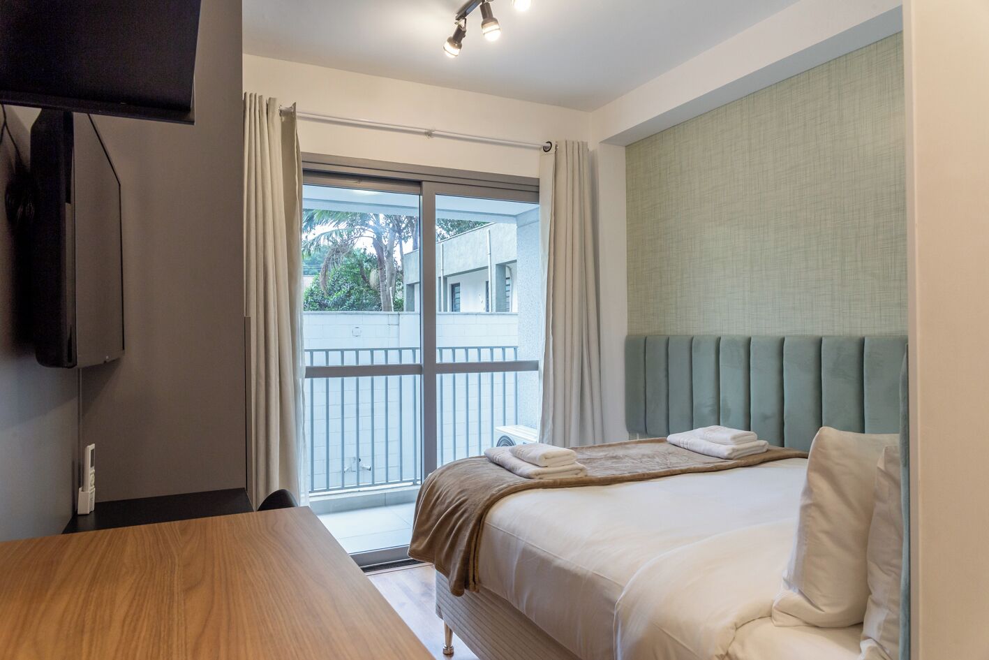 A hotel room with a bed, a TV, a wooden table, and a balcony with a view of buildings and trees.