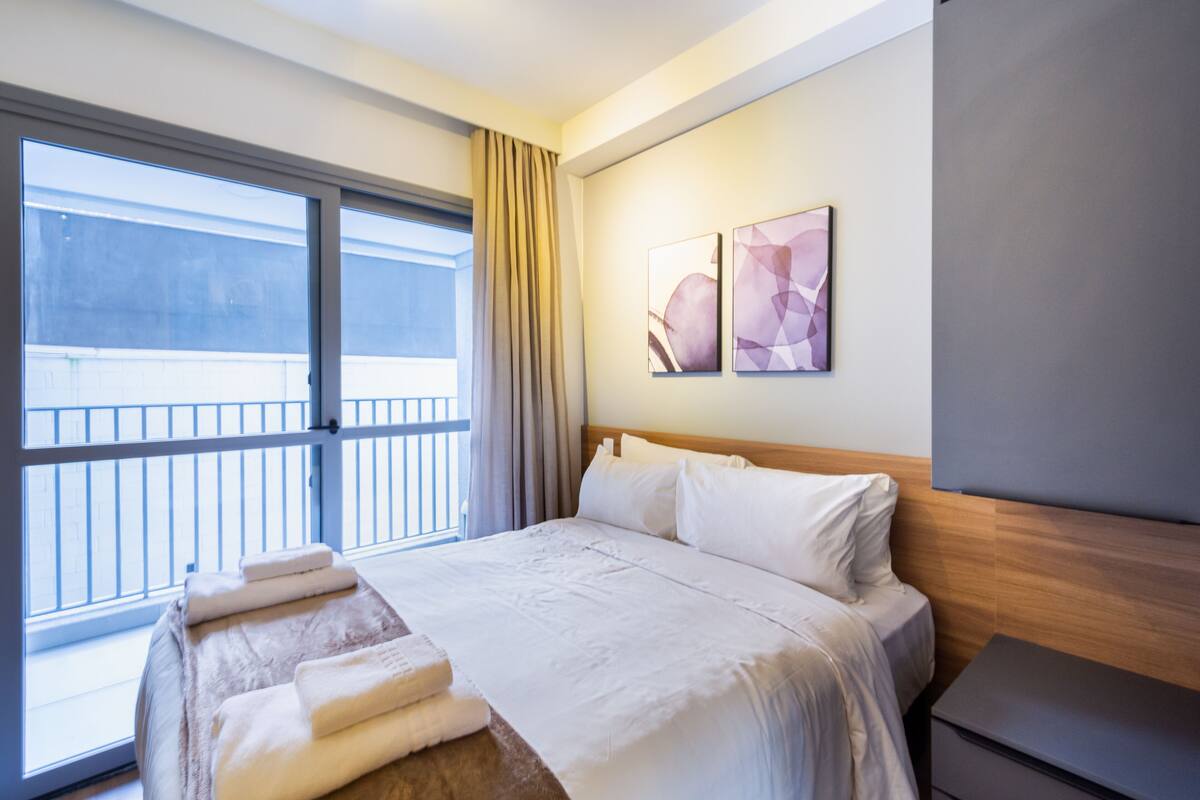Ein Hotelzimmer mit einem großen Bett, einem hölzernen Kopfteil, einem Nachttisch und einem Fenster mit Blick auf ein Gebäude.