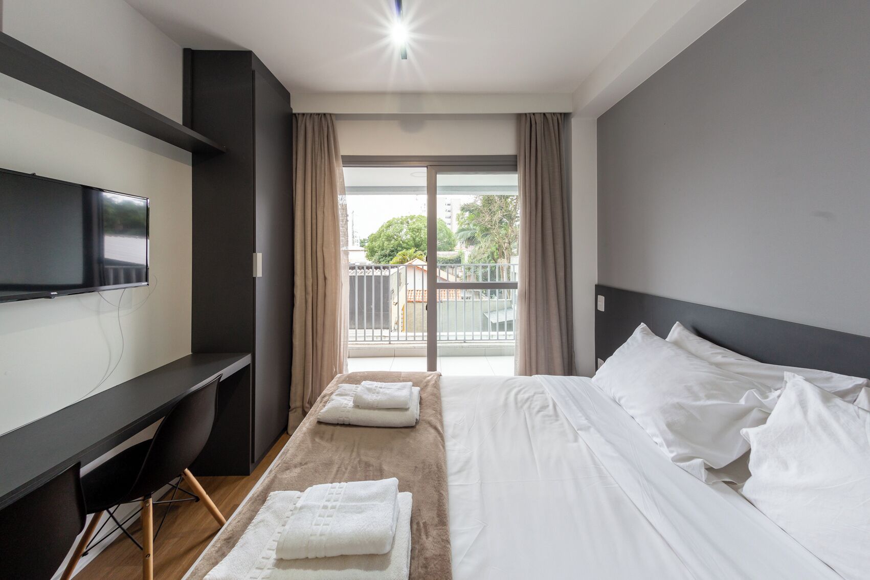 A modern hotel room with a bed, a flat-screen TV mounted on the wall, a wooden floor, and a balcony with curtains.