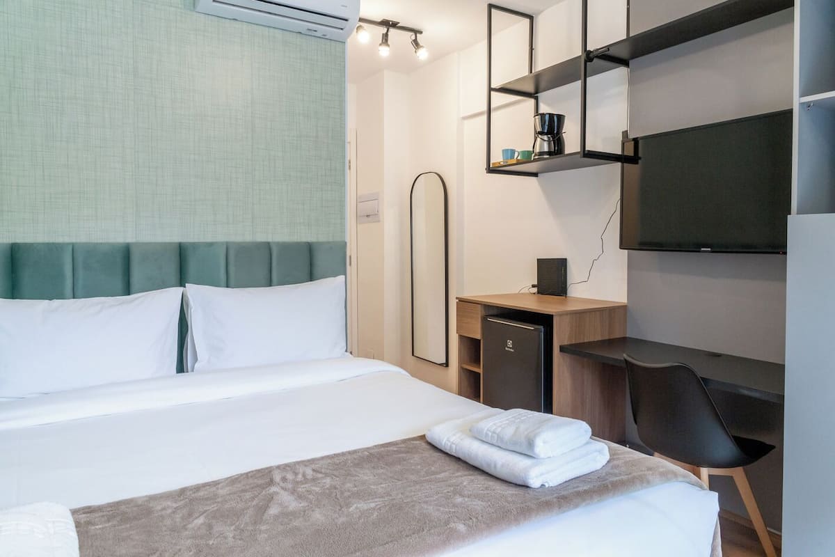 A hotel room with a bed, a desk, a chair, a TV, and a shelf with a coffee maker.