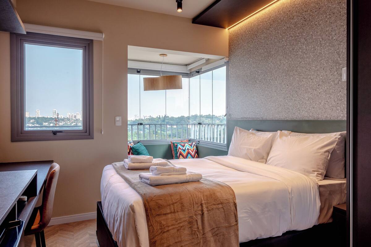Ein Hotelzimmer mit großem Fenster, einem Bett mit weißer Bettwäsche, einem Schreibtisch und einem Stuhl.