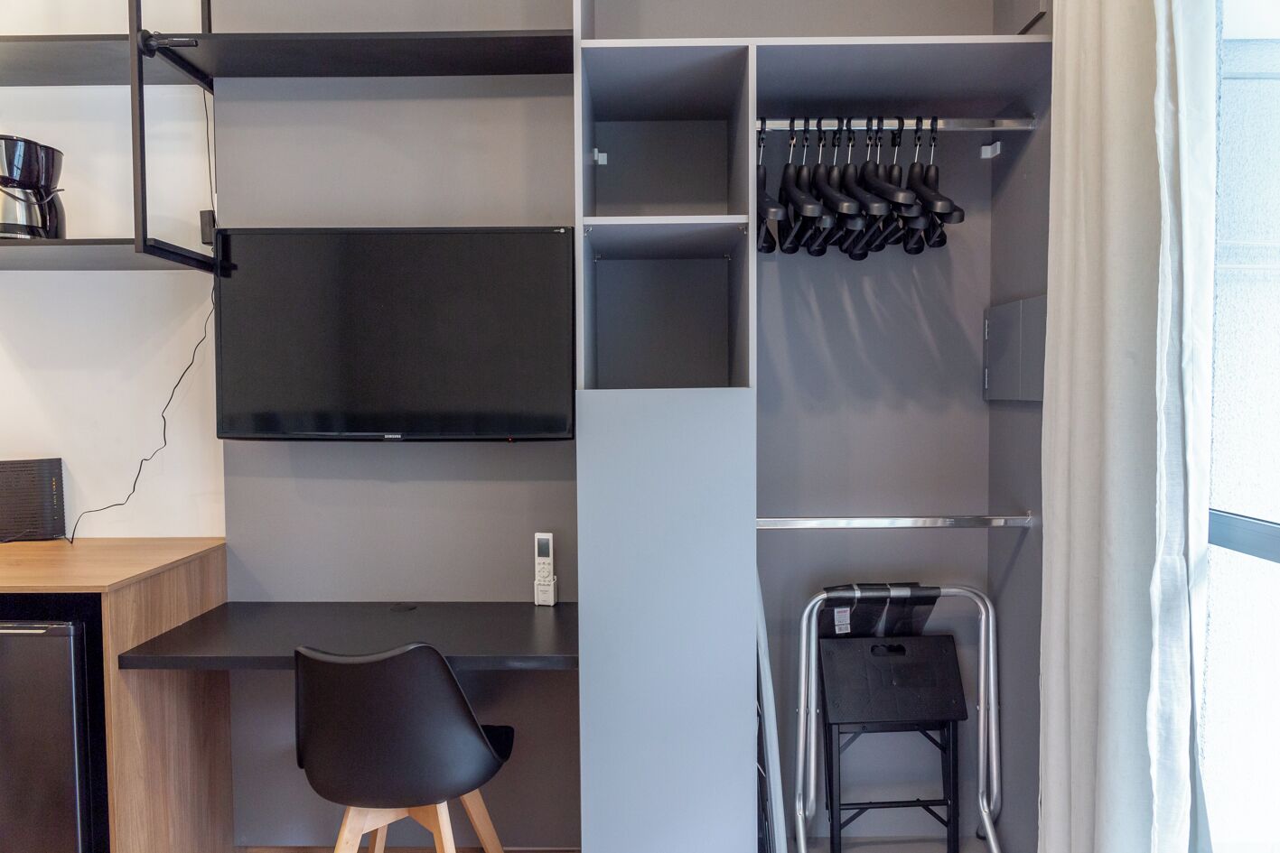 Ein modernes, aufgeräumtes Zimmer mit integriertem Fernseher, Schreibtisch mit Stuhl und Kleiderstange mit Schuhen.