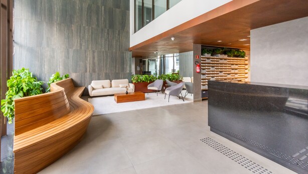 A modern lobby with a curved wooden reception desk, a sofa, and chairs. There are potted plants and a large black screen. - B.Homy - Be Urban Brooklin (São Paulo)