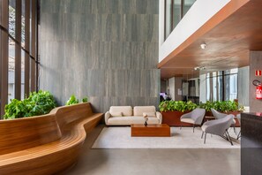 Um lobby moderno com um banco de madeira curvo, um sofá e cadeiras. Há plantas em vasos e um extintor de incêndio na parede.