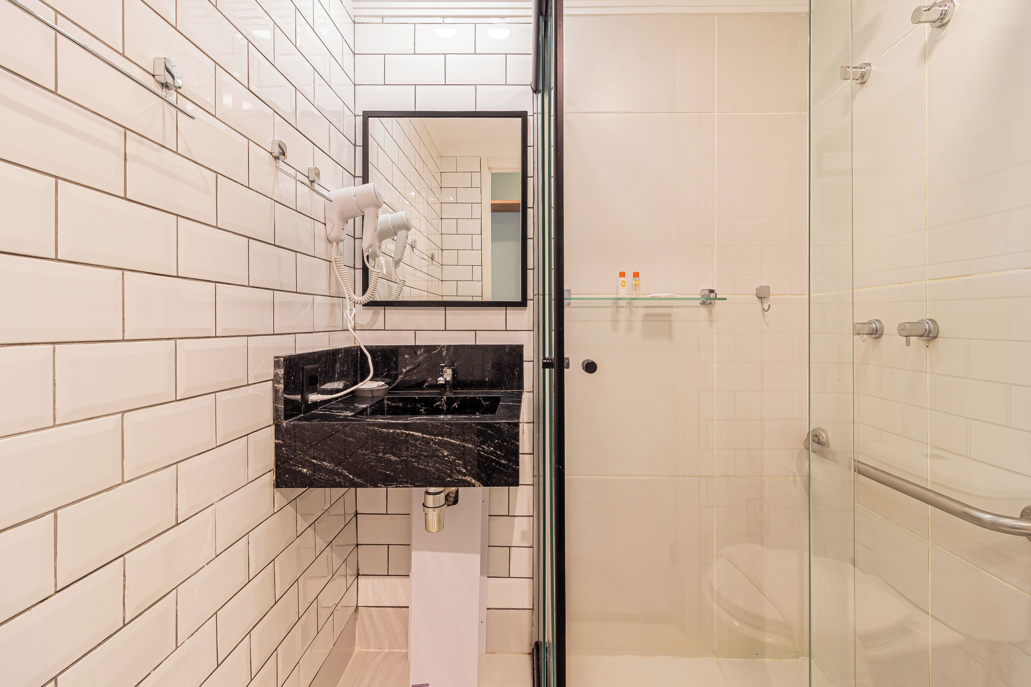 A modern bathroom with white tiled walls, a black marble sink, a handheld hair dryer, and a glass shower enclosure.