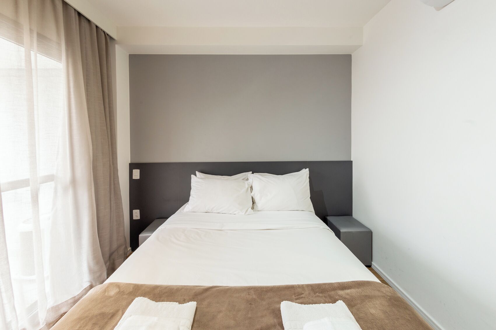 A hotel room with a bed, a nightstand, a window with curtains, and a brown rug.
