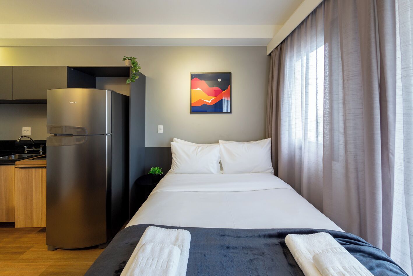 A modern hotel room with a bed, a refrigerator, a small kitchen area, and a painting on the wall.