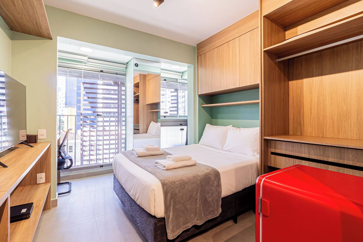 Ein Hotelzimmer mit Bett, Schreibtisch, rotem Koffer und Blick ins Freie.