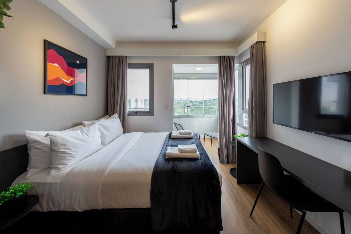 Ein modernes Hotelzimmer mit einem großen Bett, einem Schreibtisch, einem Flachbildfernseher und Blick auf die Stadt durch die Fenster.