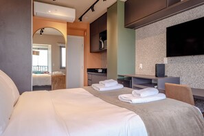 Quarto de hotel moderno com uma cama grande, uma mesa de trabalho com televisão de tela plana e uma cozinha compacta.