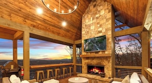 A rustic interior with a stone fireplace, wooden beams, a TV mounted on the wall, and a view of the landscape.