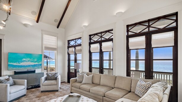A spacious living room with a large sectional sofa, two armchairs, a coffee table, and a flat-screen TV mounted on the wall. The room features large windows with dark frames and white blinds, offering a view of the ocean.
