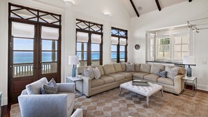 A spacious living room with a large beige sectional sofa, a smaller grey sofa, and a white ottoman. There are two side tables with lamps, and a large window with a view of the ocean.