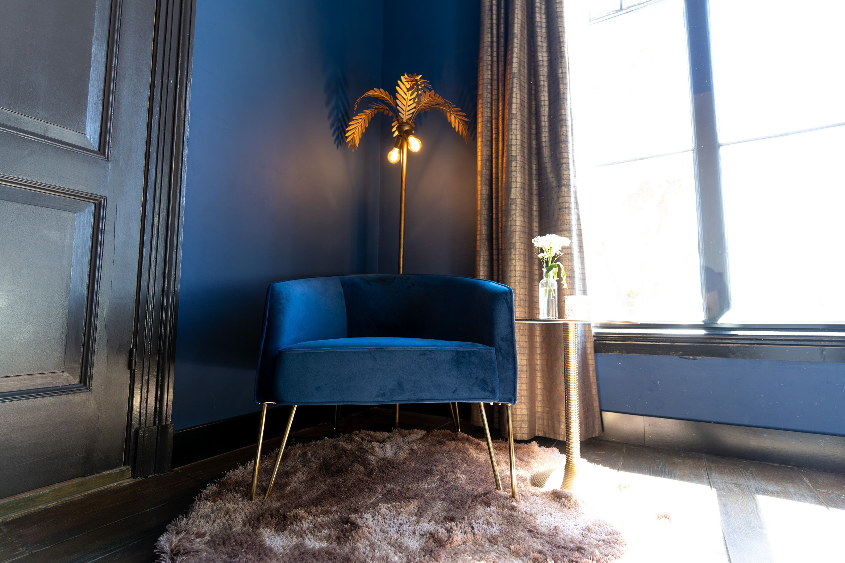 A room with a blue chair, a gold-legged floor lamp, and a window with curtains.