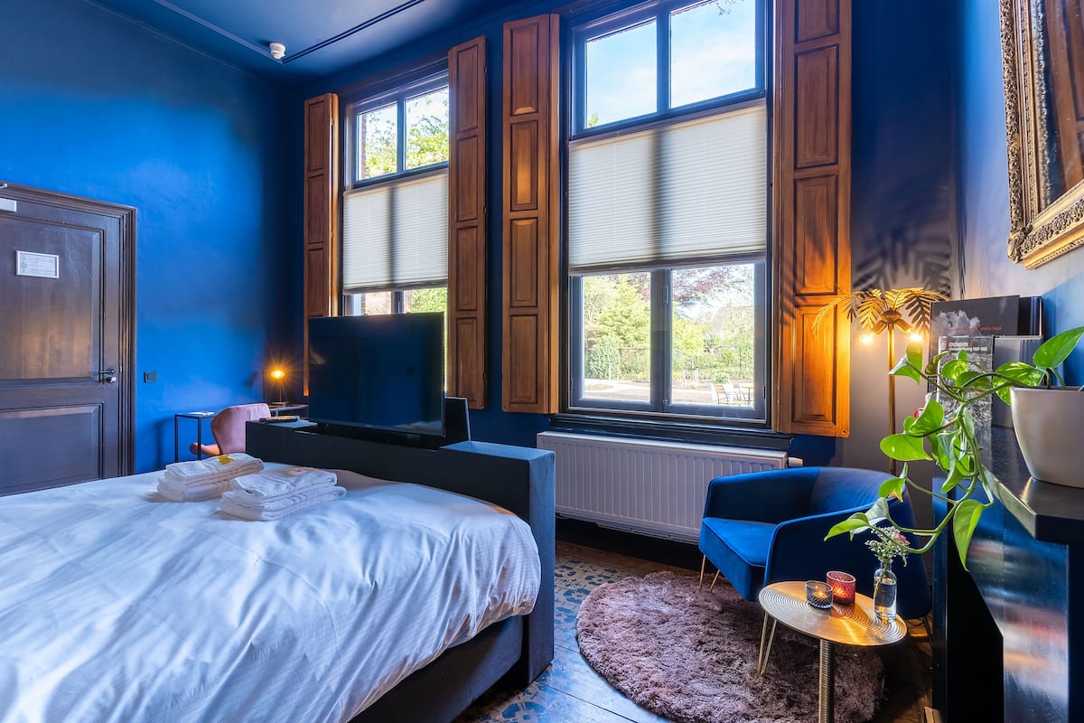 Un dormitorio con una cama, televisor, una silla, una planta y una ventana con persianas.