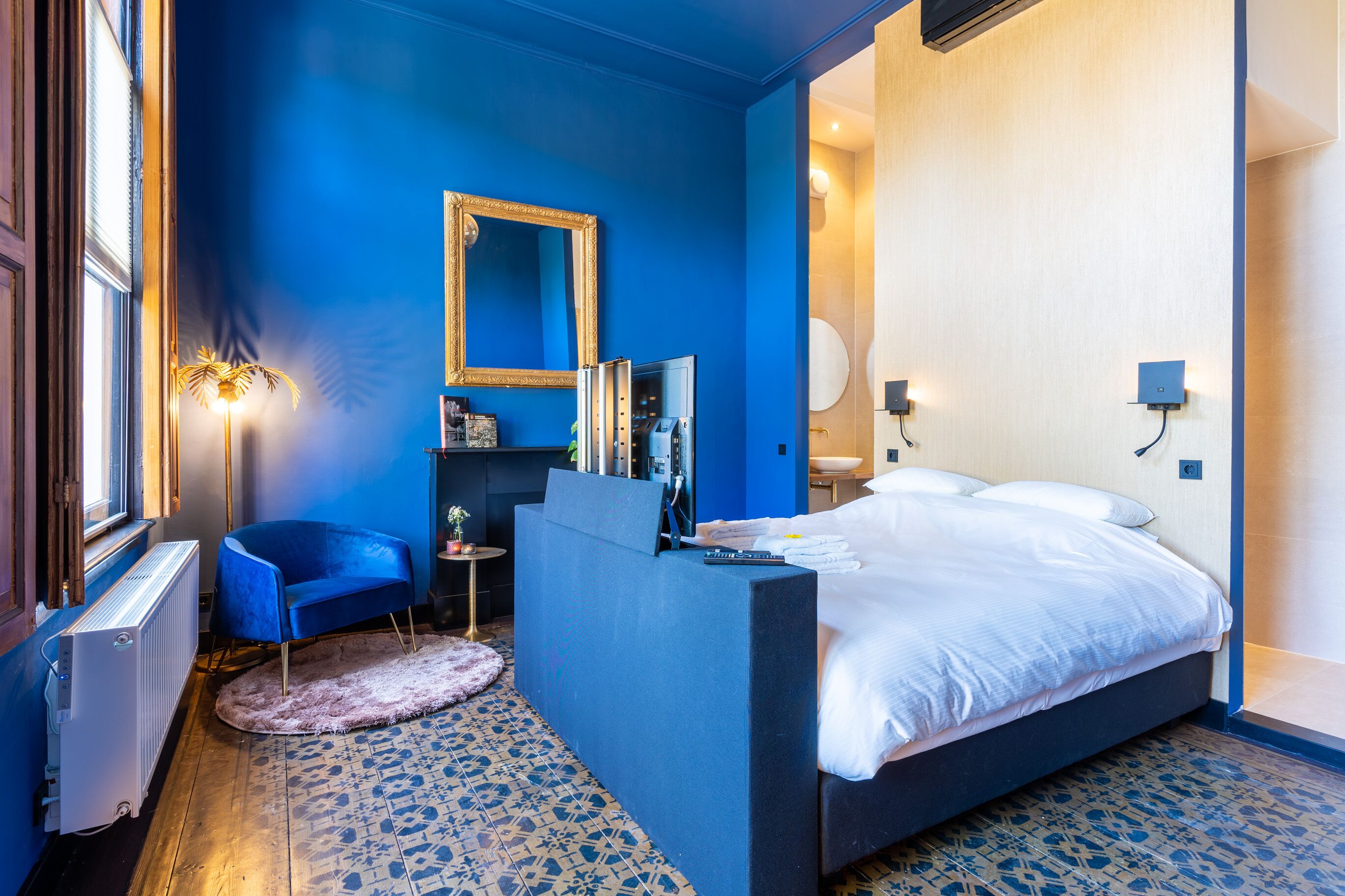 Un dormitorio moderno con una cama grande, un muro azul decorativo, un espejo, una tocador y un pequeño rincón de estar.