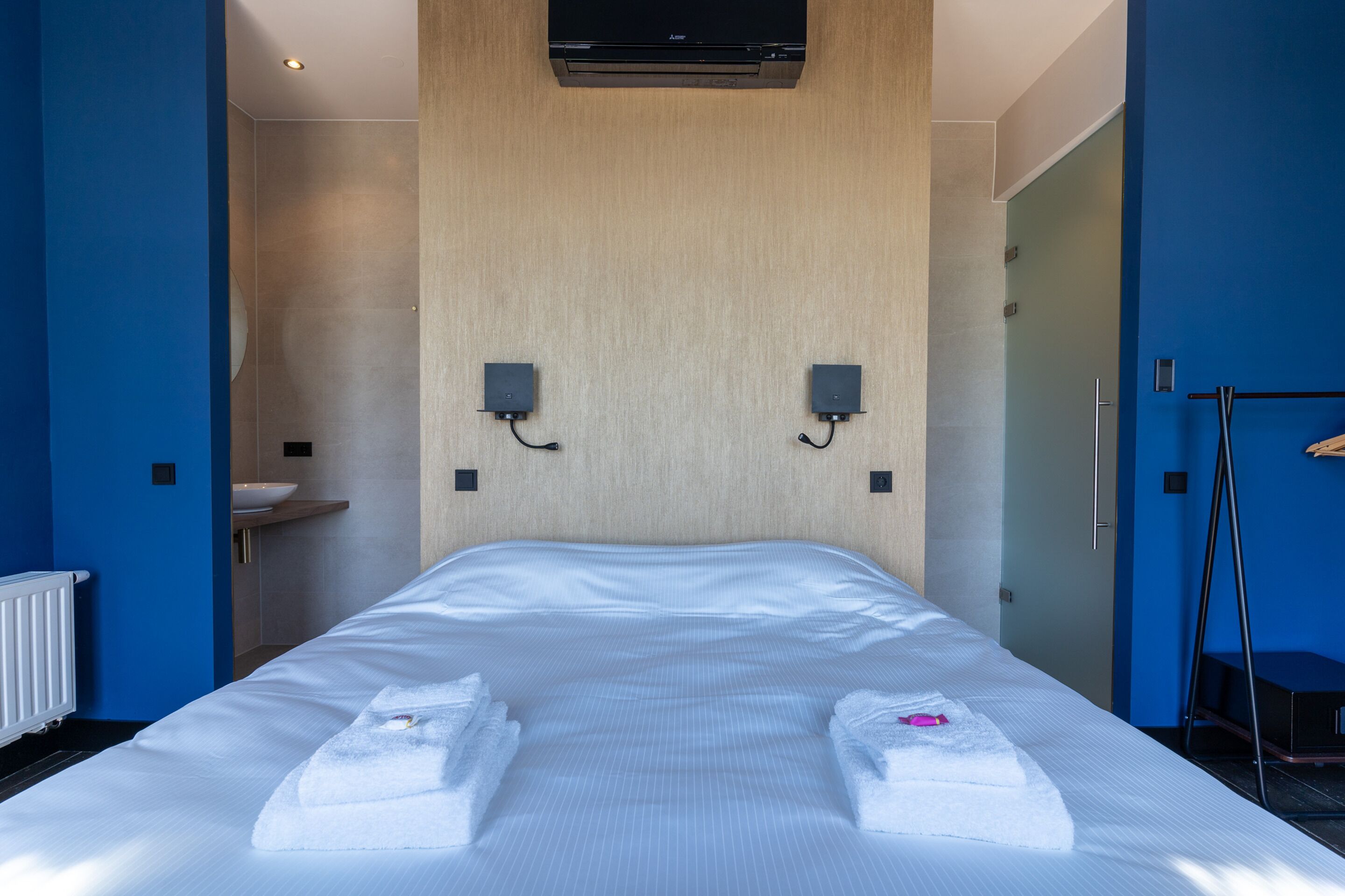 Una habitación de hotel moderna con una cama grande, dos altavoces montados en la pared, un radiador y una pared azul.