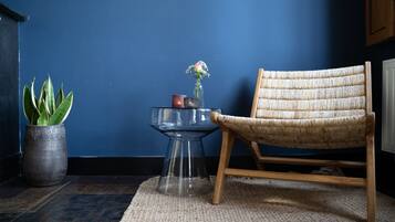 A room with a dark blue wall, a woven chair, a glass table with a vase and a potted plant.