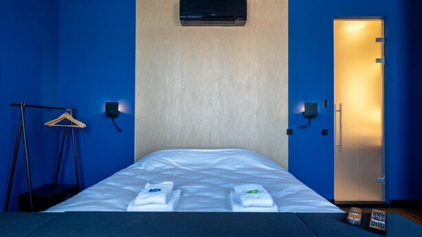 Habitación de hotel con cama, toallas, aire acondicionado montado en la pared, una puerta y una pequeña mesa con un control remoto.