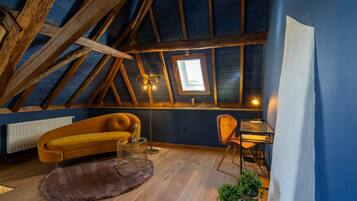 Quarto em estilo loft com teto inclinado, vigas de madeira, claraboia, sofá amarelo, mesinha, cadeira e radiador.