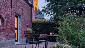 Un bâtiment en briques surmonté d’une tour horlogée, un jardin fleuri et une table entourée de chaises.
