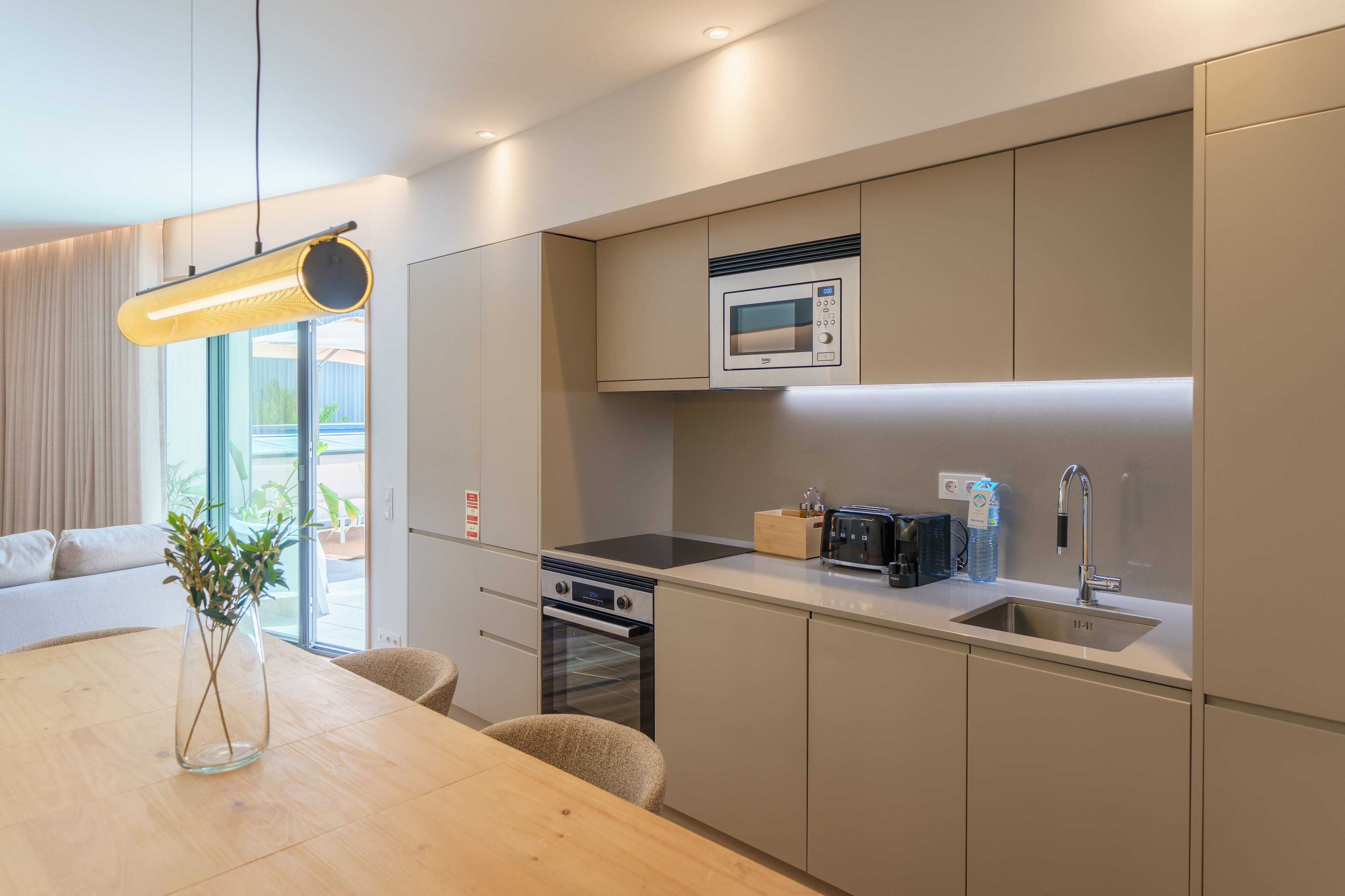 A modern kitchen with a built-in microwave, oven, and sink. There is a dining table with chairs and a vase of plants.