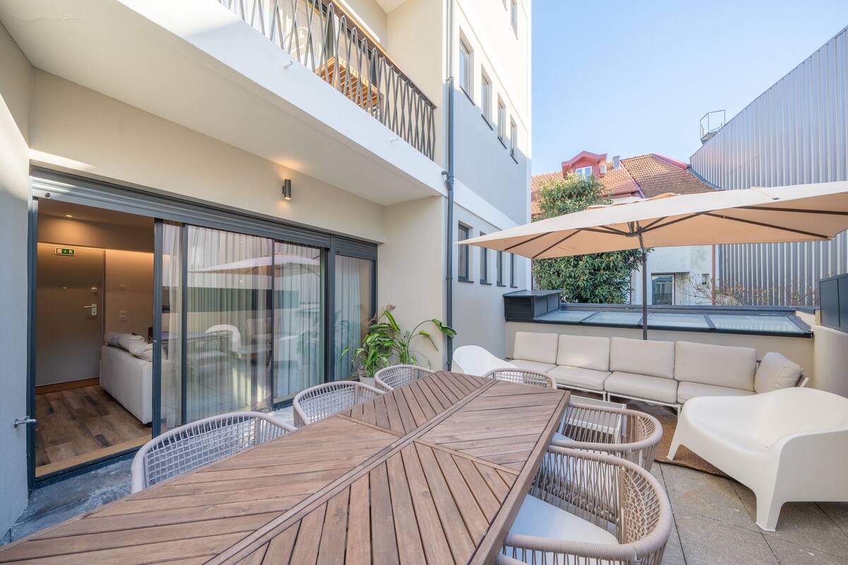 A modern outdoor seating area with a wooden table and wicker chairs, a white sofa, and a large umbrella providing shade.