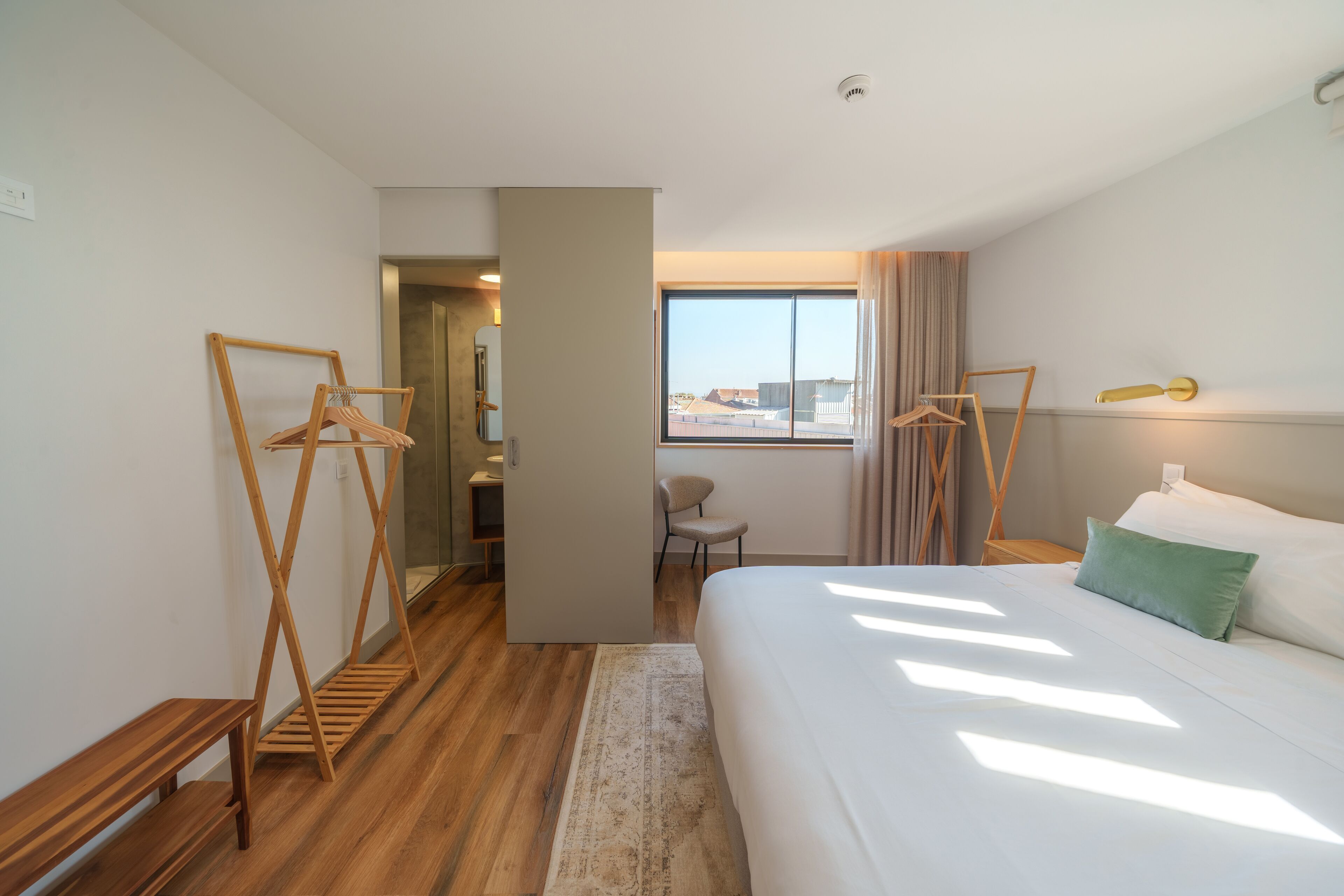 Een hotelkamer met een groot bed, een houten bankje, een kledingrek en uitzicht naar buiten door een raam.