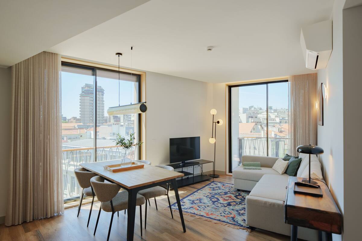 A modern living room with a dining area, large windows, a flat-screen TV, and a sofa.
