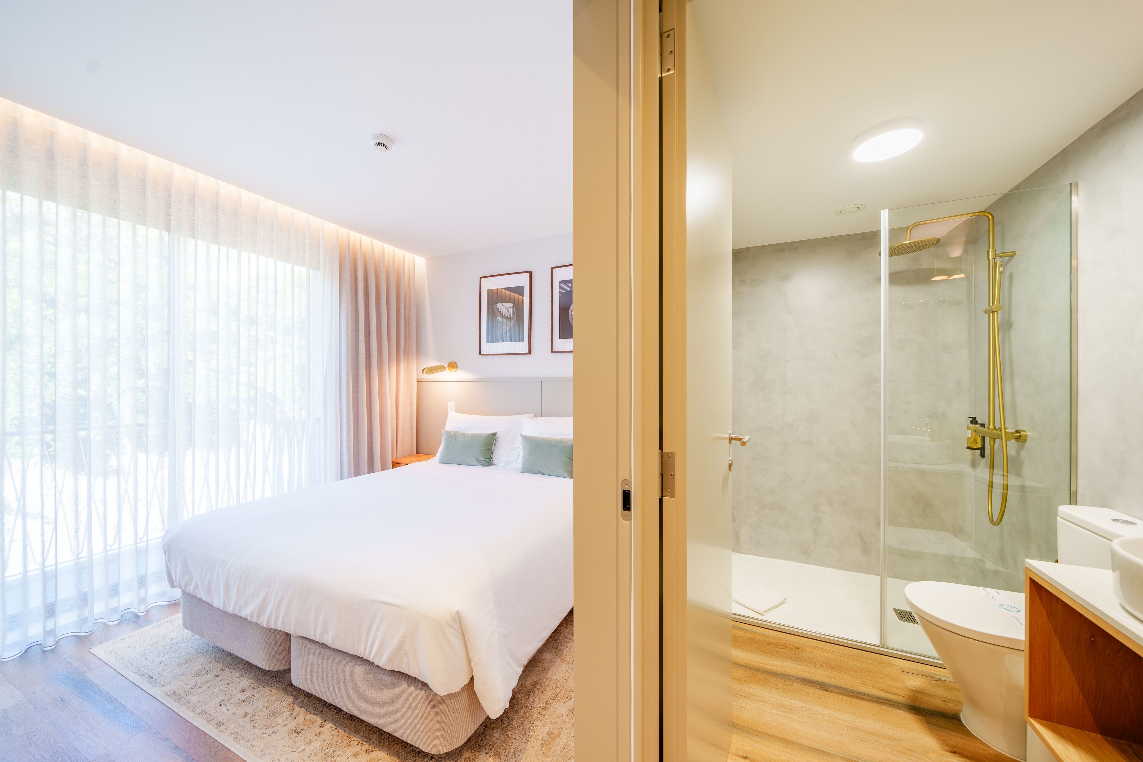 A bedroom with a large bed, a bedside table, and a window with curtains. The image also shows a bathroom with a glass shower enclosure, a toilet, and a sink.