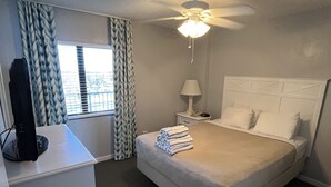 A hotel room with a bed, a desk with a computer, a nightstand with a lamp, and a ceiling fan.