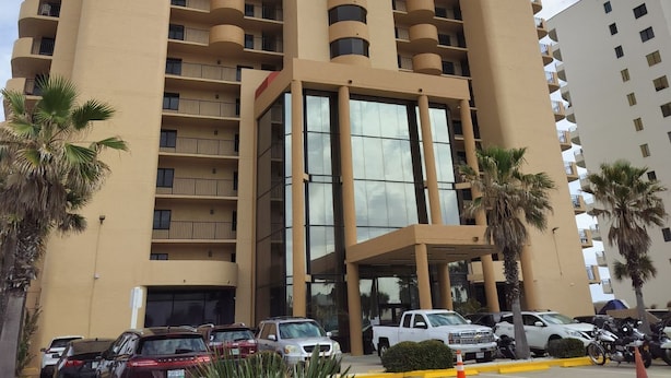 A multi-story hotel with a central glass facade, multiple balconies, and a parking area with vehicles and palm trees.