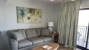 A living room with a grey sofa, a wooden coffee table, a painting of palm trees, a lamp, and a striped curtain.