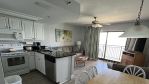 A compact kitchen with white cabinets, a stove, and a dishwasher. There's a dining area with a wooden table and chairs, a sofa, and a ceiling fan.