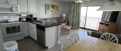 A compact kitchen with white cabinets, a stove, and a dishwasher. There's a dining area with a wooden table and chairs, a sofa, and a ceiling fan.
