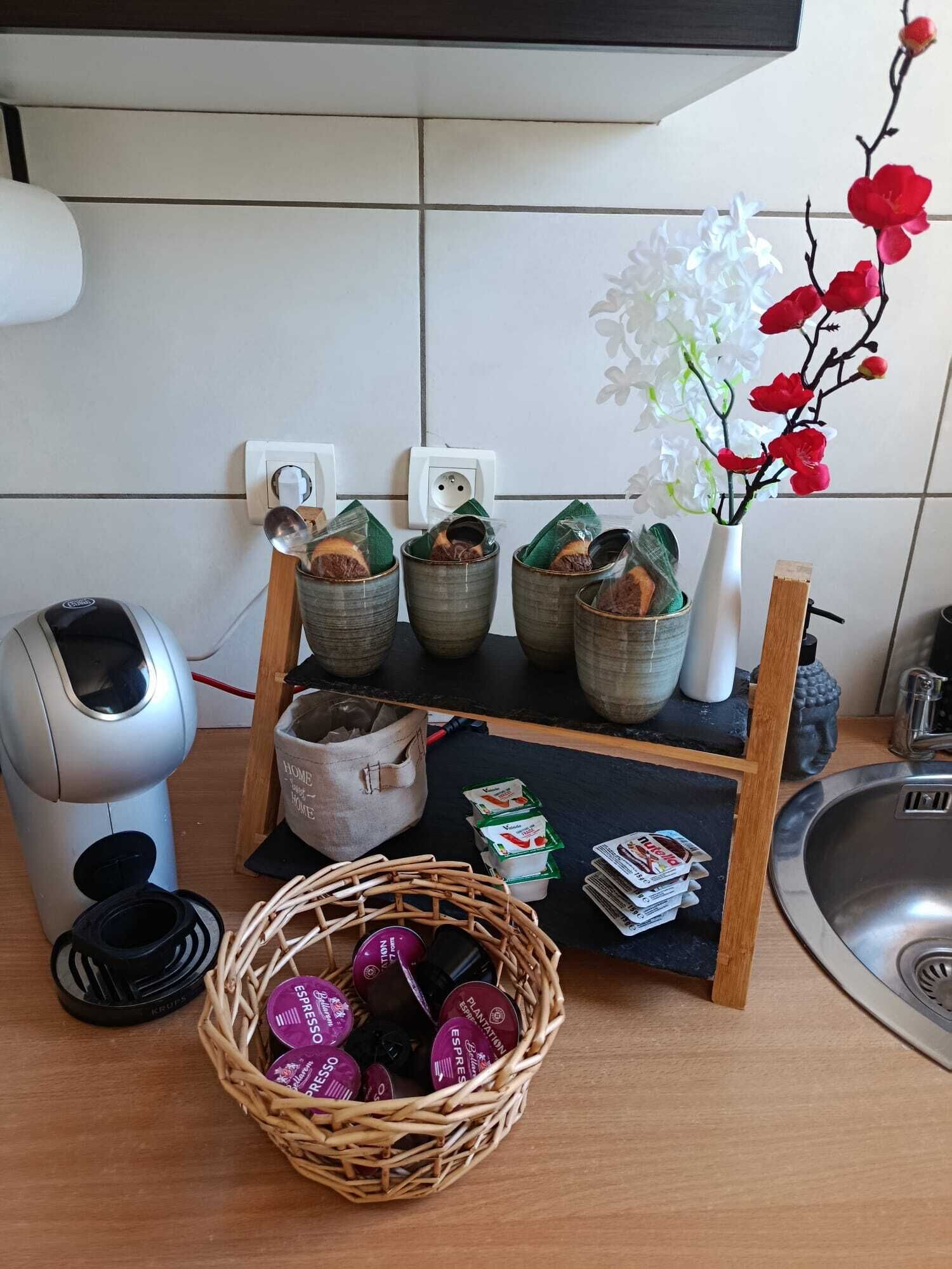 A kitchen counter with a coffee machine, a basket of coffee pods, a vase with flowers, and a small shelf with jars.