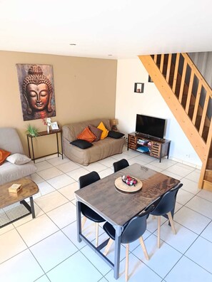 Uma sala de estar com sofá, uma mesa de jantar com cadeiras, uma televisão e uma escada.