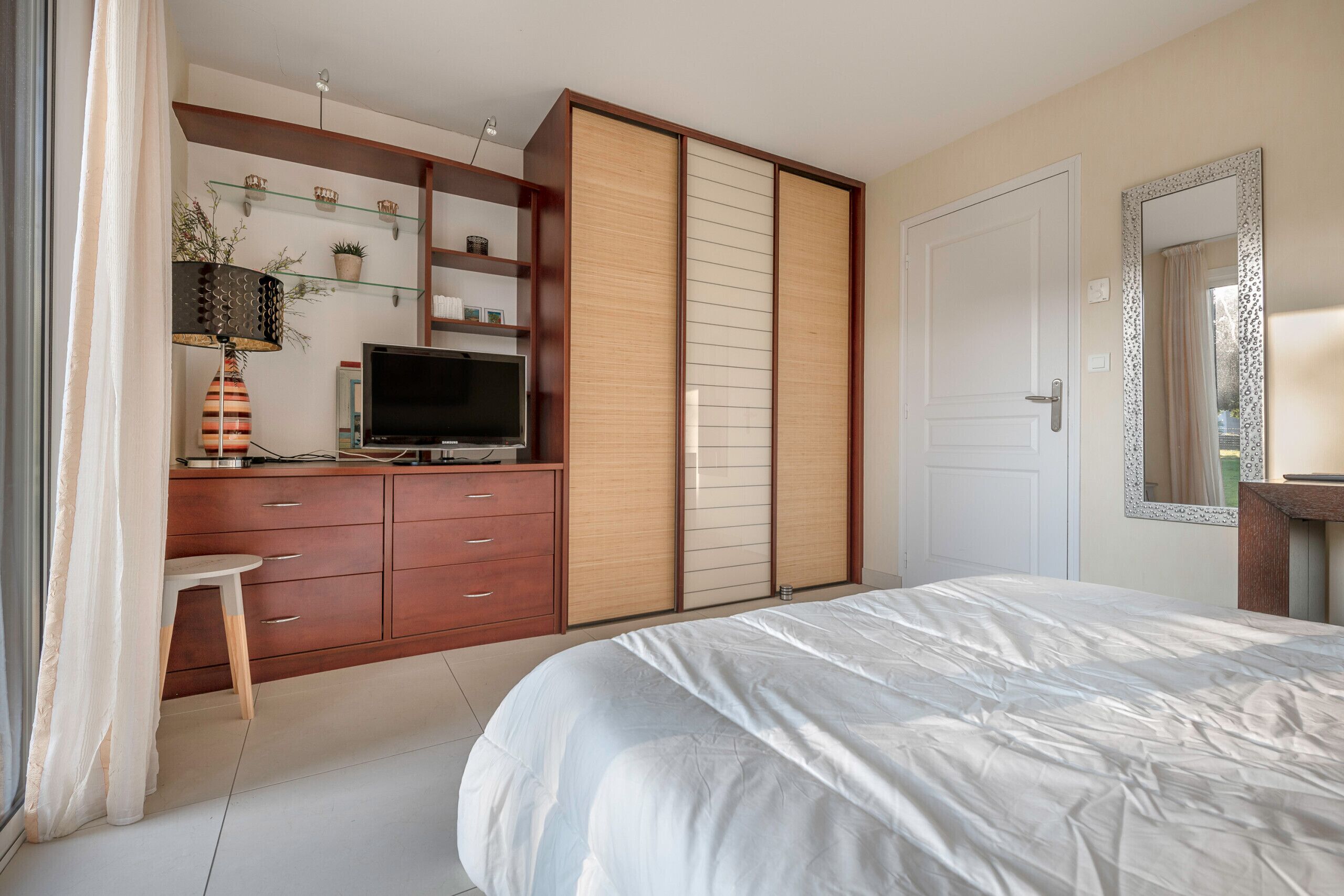 Ein Schlafzimmer mit einem Bett, einem Fernseher, einem Schreibtisch mit Schubladen, einem Regal mit Pflanzen und einem Kleiderschrank.