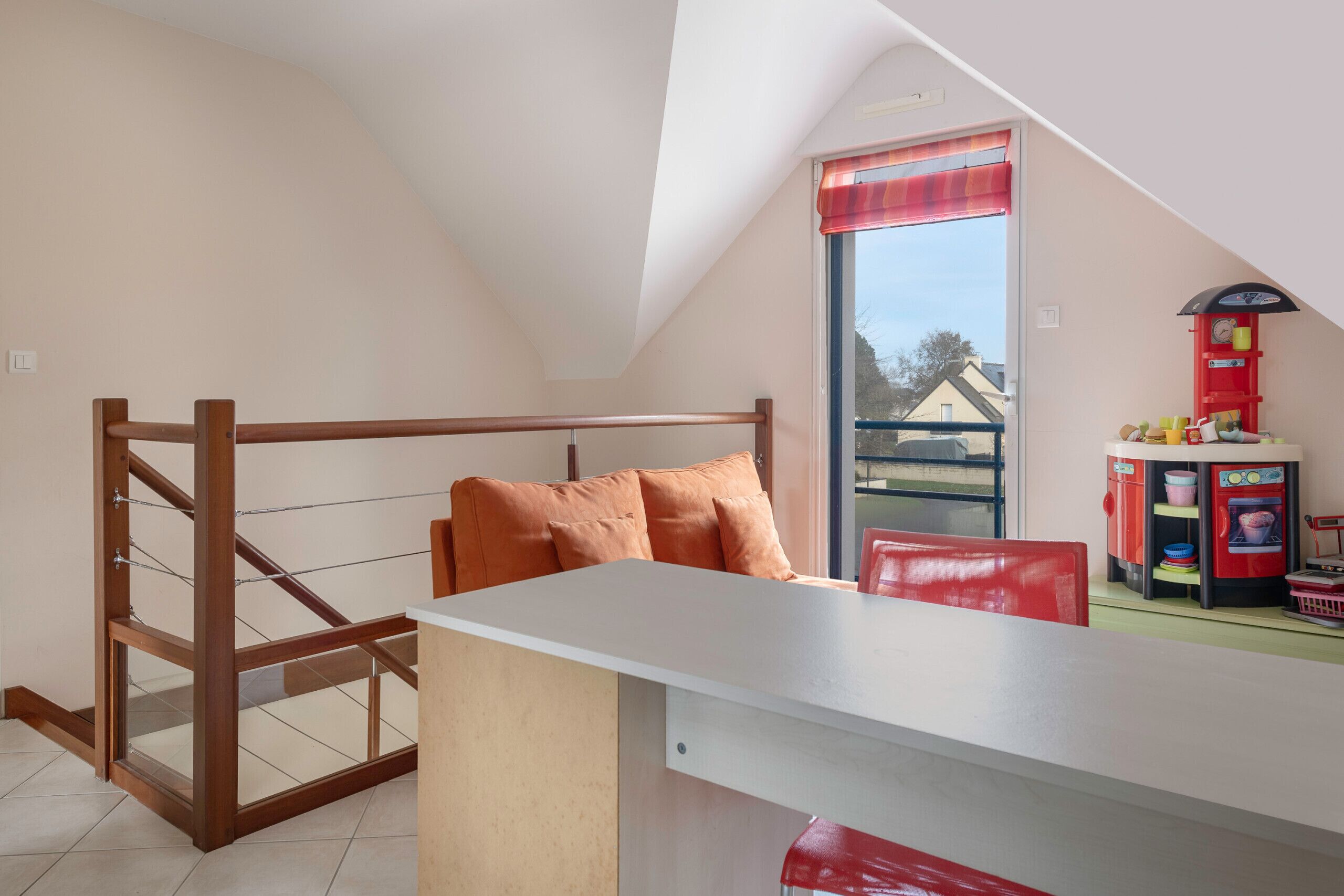 Ein Schlafzimmer im Loft-Stil mit schrägem Dach, einer Holzleiter, einem weißen Schreibtisch, einem roten Stuhl und einer roten Küchenzeile mit einer Uhr.