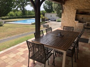 An outdoor patio with a wooden table and chairs, a stone wall, and a view of a swimming pool. - Maison Périgourdine Restaurée (Beaumontois-en-Périgord)