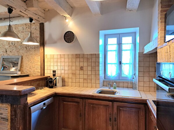 Une cuisine avec des meubles de rangement en bois, un pare-feu en carrelage, une horloge, des luminaires suspendus et une fenêtre avec des rideaux.