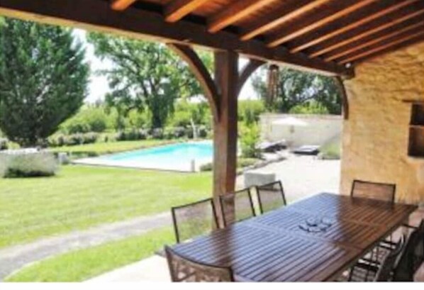 A covered patio with a wooden table and chairs, overlooking a swimming pool and garden. - Maison Périgourdine Restaurée (Beaumontois-en-Périgord)