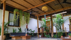 Un vaste espace de vie doté d’un plafond en bois, d’un ventilateur de plafond et de plantes suspendues. On y trouve un canapé, une table basse et une bibliothèque remplie de livres.
