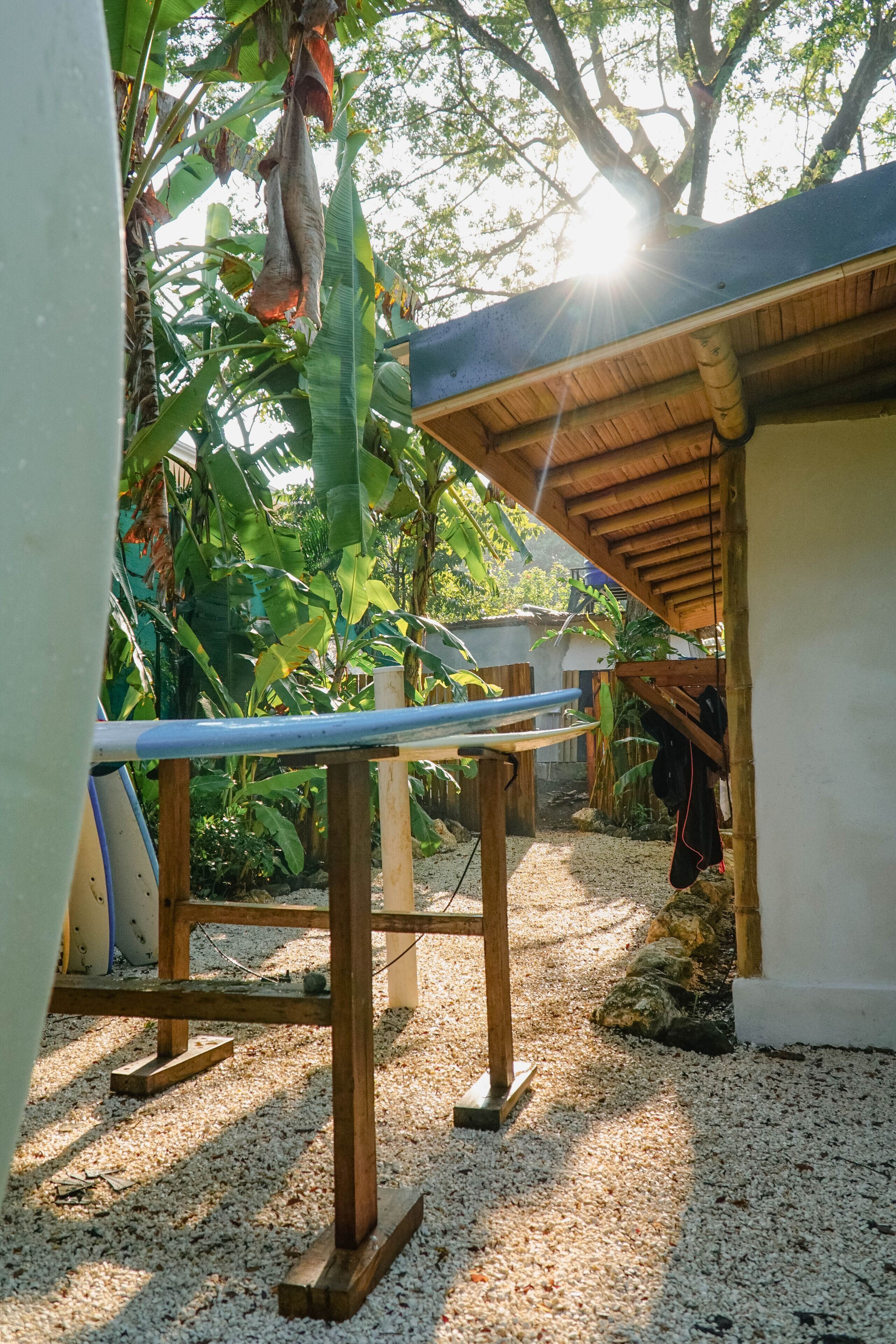 A covered outdoor area with a wooden table, surrounded by gravel and greenery.