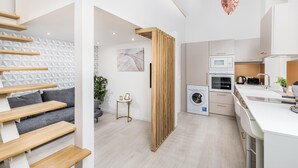 A modern kitchen with a washing machine, oven, and microwave. There is a wooden staircase and a grey sofa in the living area. - Lovely Apartment by National Museum (Budapest)
