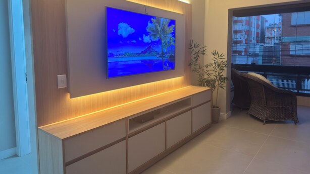 A modern living room with a flat-screen TV mounted on a wooden wall, a console table, a potted plant, wicker furniture, and a view of buildings through a window.