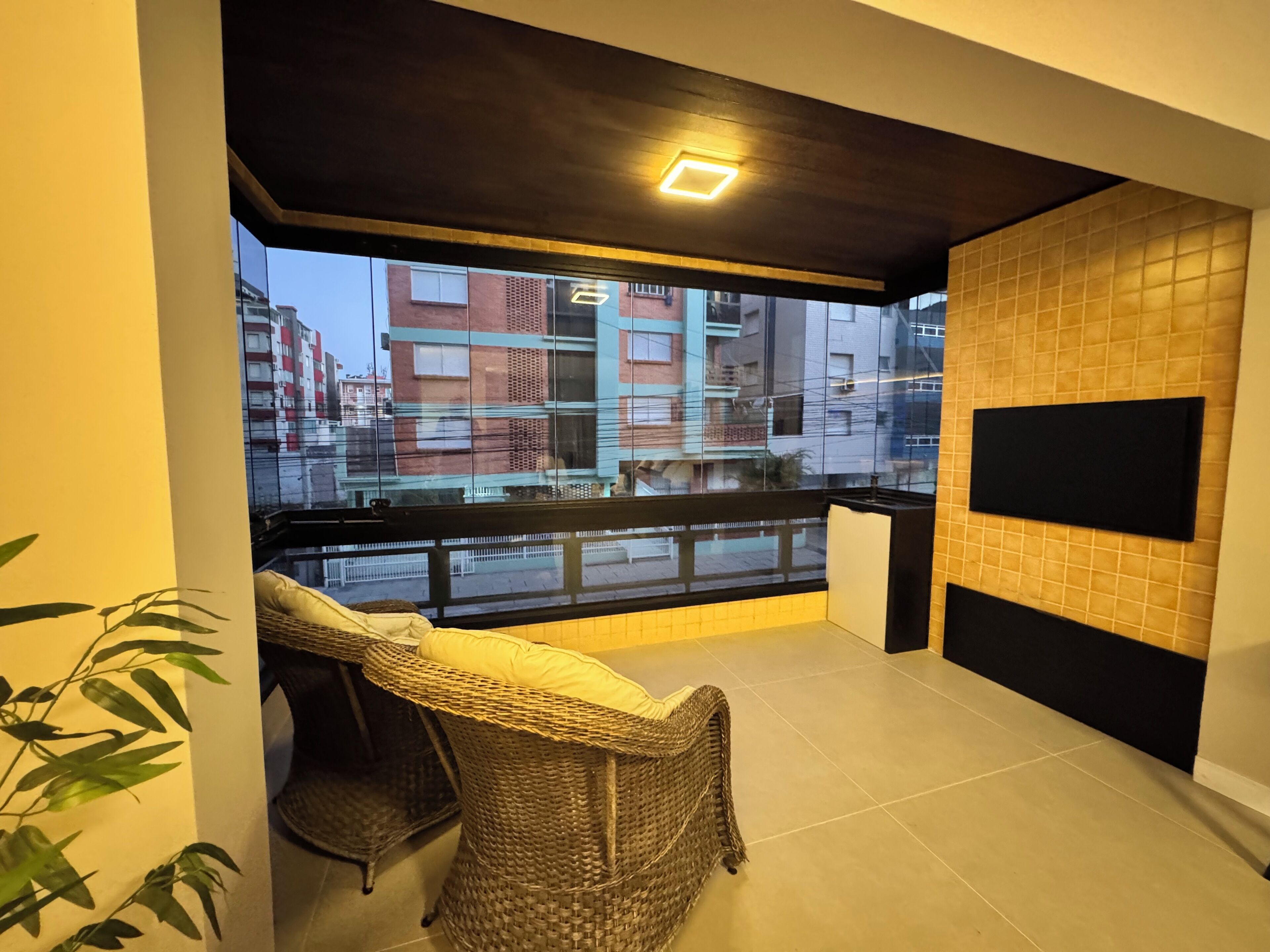 A balcony with wicker furniture, a flat-screen TV mounted on the wall, and a view of buildings in the background.