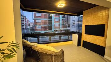 A balcony with wicker furniture, a flat-screen TV mounted on the wall, and a view of buildings in the background.